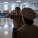 A military recruit wearing a navy blue cap labeled 'RECRUIT' is saluting a uniformed instructor or officer inside what appears to be a military training facility. Other recruits in physical training attire are visible standing in the background. The scene depicts a formal military training interaction, likely at a boot camp or basic training facility.