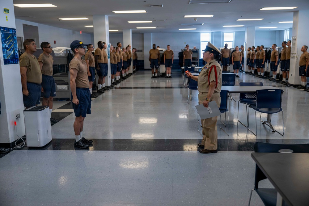 A military or naval training facility showing what appears to be an inspection or formation drill in a large barracks bay. A senior officer or petty officer in a formal uniform with a distinctive cover and red aiguillette is addressing a recruit or junior member standing at attention. Rows of personnel in tan shirts and navy blue shorts are lined up along both sides of the bay.