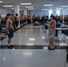 A military or naval training facility showing what appears to be an inspection or formation drill in a large barracks bay. A senior officer or petty officer in a formal uniform with a distinctive cover and red aiguillette is addressing a recruit or junior member standing at attention. Rows of personnel in tan shirts and navy blue shorts are lined up along both sides of the bay.