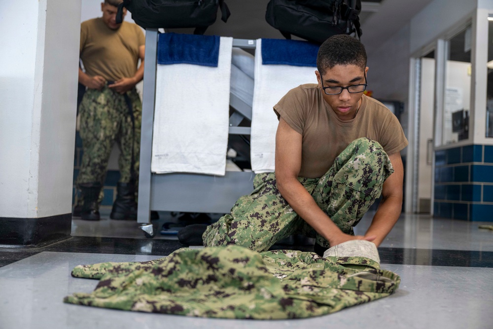 A young military service member wearing NWU (Navy Working Uniform) camouflage pants and a tan t-shirt sits cross-legged on a floor, folding or organizing camouflage uniform items. A second service member in similar uniform stands in the background near a rack with white and blue towels. The setting appears to be a military barracks or berthing area.