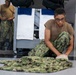 A young military service member wearing NWU (Navy Working Uniform) camouflage pants and a tan t-shirt sits cross-legged on a floor, folding or organizing camouflage uniform items. A second service member in similar uniform stands in the background near a rack with white and blue towels. The setting appears to be a military barracks or berthing area.