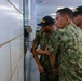 A group of military recruits in U.S. Navy digital camouflage uniforms (NWU Type III) are gathered closely together, leaning toward a wall or window in what appears to be an indoor facility. They are wearing black caps with 'RECRUIT' printed in yellow letters, indicating they are in basic training. The recruits appear to be observing or inspecting something on or through the wall surface.