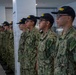 A line of U.S. Navy recruits standing at attention in an indoor facility, wearing Navy Working Uniform (NWU) Type III camouflage and black caps labeled 'RECRUIT' in yellow lettering. The recruits appear to be in a barracks or processing facility, standing in a disciplined formation. The image likely depicts a scene from Navy basic training (boot camp).