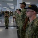 A group of U.S. Navy recruits stand at attention in what appears to be a large indoor facility, likely a recruit training command building. The recruits are wearing Navy Working Uniform (NWU Type III) camouflage and black caps with 'RECRUIT' printed in gold letters. An instructor or petty officer appears to be reviewing a tablet or clipboard while overseeing the formation.