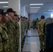 A group of U.S. Navy recruits in camouflage uniforms (NWU Type III) stand at attention in two facing rows inside what appears to be a barracks bay. Several recruits wear black caps with yellow 'REC' (Recruit) lettering. A uniformed instructor or petty officer is visible further down the aisle, suggesting a barracks inspection or formation drill.