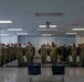 A large group of military personnel in matching camouflage uniforms and dark caps are standing at attention in two parallel rows inside what appears to be an indoor facility, possibly a training or administrative building. The formation faces inward toward a central aisle where two empty blue chairs are positioned in the foreground. The setting suggests a formal military muster, inspection, or briefing formation.
