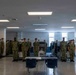 A large group of military personnel in Navy Working Uniform (NWU) camouflage stands at attention in what appears to be a military barracks or berthing area. The personnel are arranged in formation facing a central aisle, with a table and chairs positioned in the foreground. The setting suggests a formal inspection, muster, or briefing event.