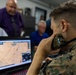 A U.S. Marine in digital camouflage (MARPAT) uniform is using a handheld military radio while monitoring a tactical mapping application on a laptop computer. Several civilians in the background appear to be observing the operation, suggesting this may be a demonstration or briefing event. The setting appears to be an indoor operations or command center environment.
