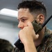 A military service member in digital camouflage (MARPAT) uniform is holding a Motorola radio/walkie-talkie to his ear, appearing to be actively listening or communicating. The setting appears to be an indoor facility with fluorescent lighting. A second uniformed individual is partially visible in the background.