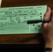 A hand holding a pen is filling out a military Artillery Fire Support Card (Green), a standardized US Army form used to coordinate and record artillery fire missions. The card contains pre-printed fields for target location, target description, method of engagement, and battle damage assessment. The card is resting on what appears to be a notebook on a wooden surface.