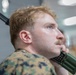 A young male service member in U.S. Marine Corps MARPAT (woodland digital camouflage) uniform is holding a military-style radio handset to his jaw/chin area. He appears to be listening or communicating, with his gaze directed upward and to the side. The image is a close-up portrait shot taken in what appears to be an indoor environment.