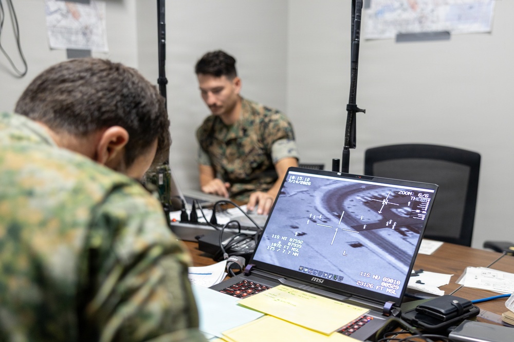 Two military personnel in camouflage uniforms are working at a table in what appears to be a tactical operations center or command post. An MSI laptop in the foreground displays what appears to be aerial surveillance or drone feed footage showing an overhead view of a location. The personnel appear to be conducting ISR (Intelligence, Surveillance, and Reconnaissance) operations or mission planning.