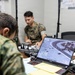 Two military personnel in camouflage uniforms are working at a table in what appears to be a tactical operations center or command post. An MSI laptop in the foreground displays what appears to be aerial surveillance or drone feed footage showing an overhead view of a location. The personnel appear to be conducting ISR (Intelligence, Surveillance, and Reconnaissance) operations or mission planning.
