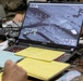 A military operations setting showing an MSI laptop displaying what appears to be aerial surveillance or drone feed footage with targeting crosshairs and telemetry data. Personnel in military camouflage uniforms are visible in the background, and the desk is covered with notes, yellow legal pads, and various cables. The screen shows coordinates, altitude readings, and a timestamp of 10:15:32 on 7/14/2026.