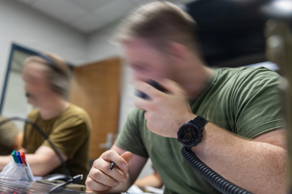 A young man wearing a green t-shirt and a black digital watch is seated at a desk, holding a corded telephone handset to his face while gripping a pen in his other hand. A second individual in a tan/olive shirt is visible in the background, also seated at a desk. The foreground subject is motion-blurred, suggesting deliberate shallow depth-of-field photography.
