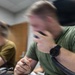 A young man wearing a green t-shirt and a black digital watch is seated at a desk, holding a corded telephone handset to his face while gripping a pen in his other hand. A second individual in a tan/olive shirt is visible in the background, also seated at a desk. The foreground subject is motion-blurred, suggesting deliberate shallow depth-of-field photography.
