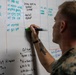A military service member in digital camouflage uniform is writing on a whiteboard with a green marker. The whiteboard contains tactical or operational information including grid coordinates, status entries, and chief's notes. The scene appears to be a military operations or briefing room setting.