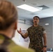 A U.S. Marine in woodland MARPAT camouflage uniform is speaking or briefing to at least one other person in what appears to be an indoor military facility or office setting. The Marine, whose name tape reads 'ARRIAGA,' is gesturing with his right hand, suggesting active communication or instruction. A second individual is visible in the foreground, blurred and out of focus, facing the speaking Marine.