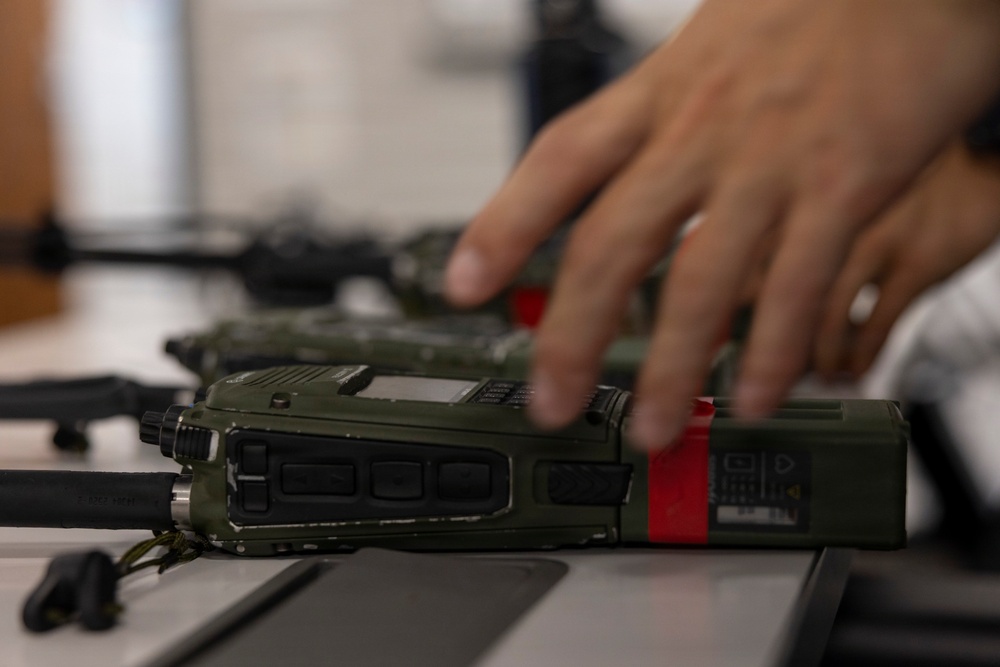 A close-up image showing military-grade handheld radio transceivers in olive drab/green coloring laid on a flat surface. A person's hand is visible reaching toward or handling one of the radios. The radios feature red tape markings, multiple buttons, a small display screen, and attached antennas.