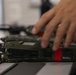 A close-up image showing military-grade handheld radio transceivers in olive drab/green coloring laid on a flat surface. A person's hand is visible reaching toward or handling one of the radios. The radios feature red tape markings, multiple buttons, a small display screen, and attached antennas.