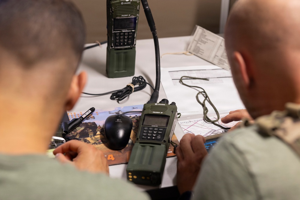 Two individuals in military-style clothing are gathered around a table examining maps and documents alongside military-grade radio equipment. The scene appears to be a tactical operations or communications setup, with L3Harris Falcon III handheld radios prominently displayed. The personnel appear to be reviewing mission planning materials or operational maps.