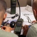 Two individuals in military-style clothing are gathered around a table examining maps and documents alongside military-grade radio equipment. The scene appears to be a tactical operations or communications setup, with L3Harris Falcon III handheld radios prominently displayed. The personnel appear to be reviewing mission planning materials or operational maps.
