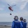 Three naval personnel in blue flight deck uniforms and cranial helmets observe a red helicopter hovering above a ship's flight deck over open ocean. The helicopter, consistent with a Coast Guard HH-65 Dolphin, is conducting a hoist or cargo transfer operation with a load suspended beneath it. This appears to be a vertical replenishment or search-and-rescue training evolution conducted at sea.