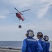 Three naval personnel in blue flight deck uniforms and cranial helmets observe a red helicopter hovering above a ship's flight deck over open ocean. The helicopter, consistent with a Coast Guard HH-65 Dolphin, is conducting a hoist or cargo transfer operation with a load suspended beneath it. This appears to be a vertical replenishment or search-and-rescue training evolution conducted at sea.