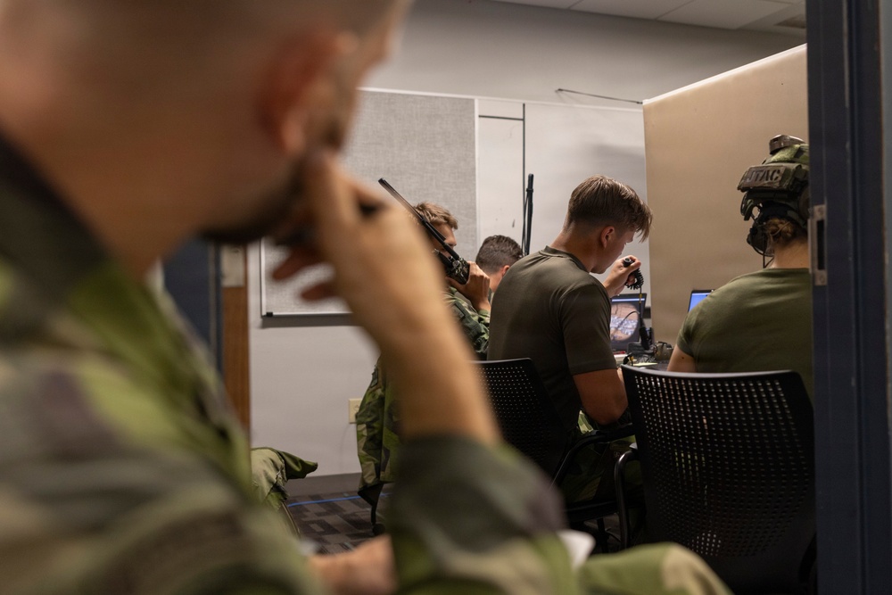 Military personnel in green/olive uniforms are gathered in what appears to be an indoor operations or training room. Several individuals are seated at workstations with computer monitors, while others are using handheld radios and communication equipment. One person on the right is wearing tactical headgear with visible 'JTAC' lettering, suggesting joint terminal attack controller training or operations.