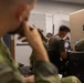 Military personnel in green/olive uniforms are gathered in what appears to be an indoor operations or training room. Several individuals are seated at workstations with computer monitors, while others are using handheld radios and communication equipment. One person on the right is wearing tactical headgear with visible 'JTAC' lettering, suggesting joint terminal attack controller training or operations.