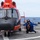 A crew member in blue flight deck gear crouches on a ship's helicopter landing pad beside a red and black helicopter, appearing to give a thumbs-up signal. The scene takes place on the flight deck of what appears to be a U.S. Coast Guard cutter, with open ocean visible in the background. A second vessel is faintly visible on the horizon.