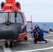 A crew member in blue flight deck gear crouches on a ship's helicopter landing pad beside a red and black helicopter, appearing to give a thumbs-up signal. The scene takes place on the flight deck of what appears to be a U.S. Coast Guard cutter, with open ocean visible in the background. A second vessel is faintly visible on the horizon.