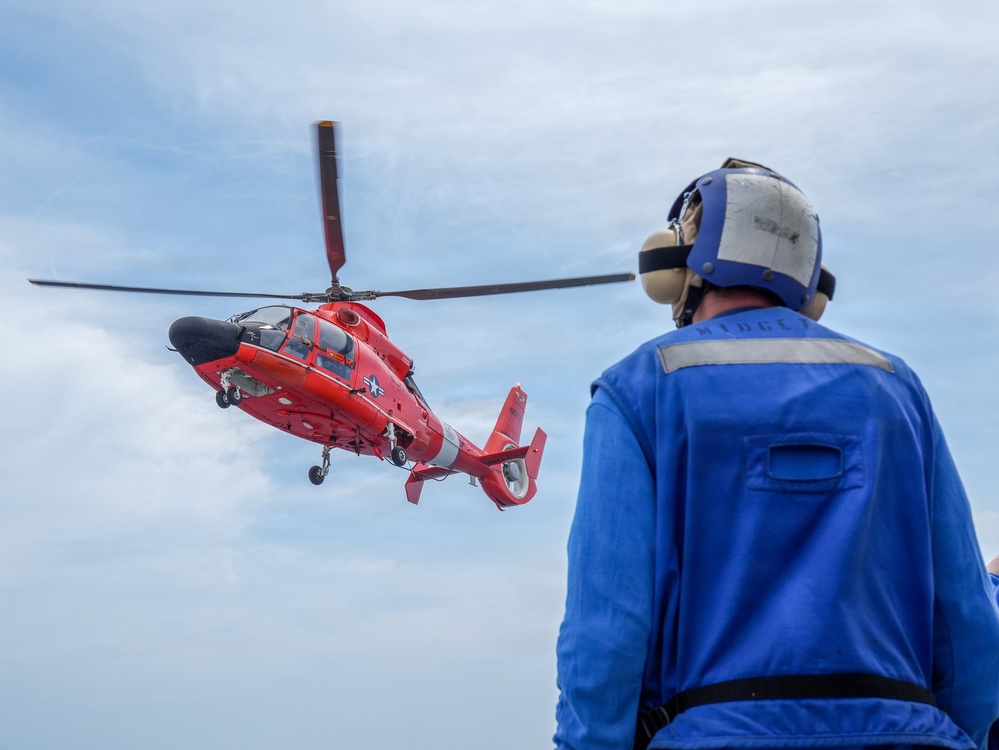 A red U.S. Coast Guard helicopter, identifiable by its distinctive red paint scheme and star insignia, hovers or approaches a landing area while a deck crew member in a blue flight suit and helmet observes from the foreground. The personnel is wearing hearing protection and appears to be a flight deck handler or landing signal officer. The scene likely takes place on a ship's flight deck or elevated platform over open water.