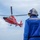 A red U.S. Coast Guard helicopter, identifiable by its distinctive red paint scheme and star insignia, hovers or approaches a landing area while a deck crew member in a blue flight suit and helmet observes from the foreground. The personnel is wearing hearing protection and appears to be a flight deck handler or landing signal officer. The scene likely takes place on a ship's flight deck or elevated platform over open water.