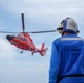 A red U.S. Coast Guard helicopter, identifiable by its distinctive red paint scheme and star insignia, hovers or approaches a landing area while a deck crew member in a blue flight suit and helmet observes from the foreground. The personnel is wearing hearing protection and appears to be a flight deck handler or landing signal officer. The scene likely takes place on a ship's flight deck or elevated platform over open water.