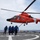 A bright orange U.S. Coast Guard helicopter (tail number 6507) hovers low over a ship's flight deck while four crew members in blue flight suits walk away from it. The helicopter appears to be an Aerospatiale HH-65 Dolphin, a standard Coast Guard rotary-wing aircraft. The scene takes place at sea, with open ocean visible in the background.