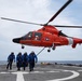 A bright orange U.S. Coast Guard helicopter (tail number 6507) hovers low over a ship's flight deck while four crew members in blue flight suits walk away from it. The helicopter appears to be an Aerospatiale HH-65 Dolphin, a standard Coast Guard rotary-wing aircraft. The scene takes place at sea, with open ocean visible in the background.