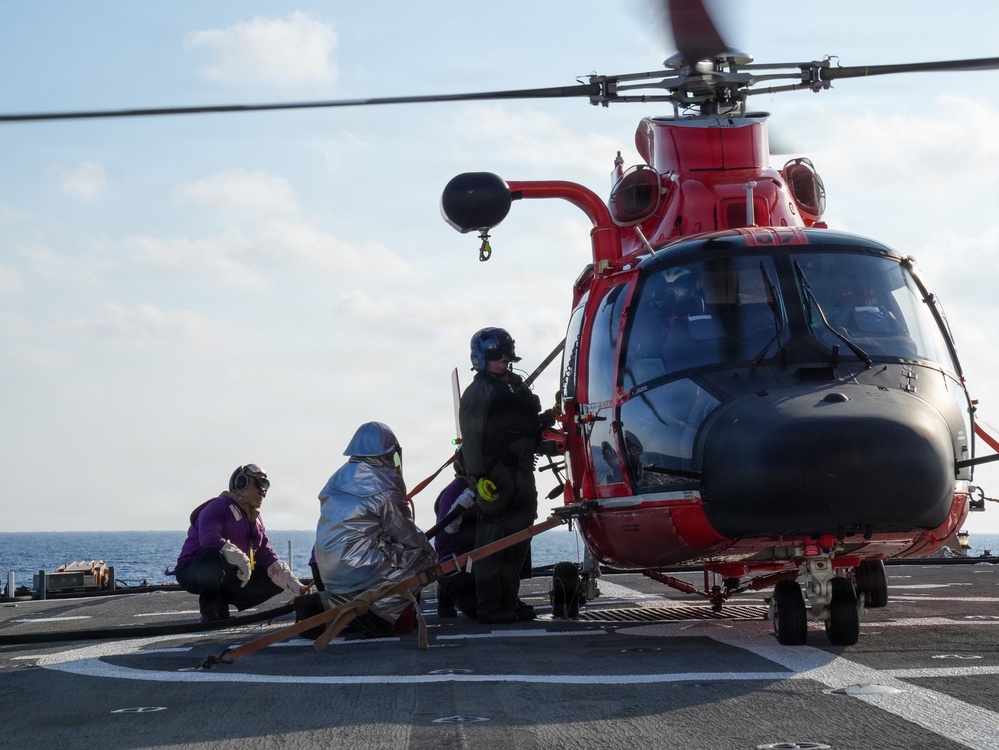 A red helicopter, consistent with a U.S. Coast Guard MH-65 Dolphin, is landed on the flight deck of a naval vessel at sea. Several personnel in specialized gear are working around the aircraft, with one individual appearing to be on a stretcher or litter, suggesting a medical evacuation or rescue operation. The rotor blades are spinning, indicating the helicopter is active.