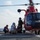 A red helicopter, consistent with a U.S. Coast Guard MH-65 Dolphin, is landed on the flight deck of a naval vessel at sea. Several personnel in specialized gear are working around the aircraft, with one individual appearing to be on a stretcher or litter, suggesting a medical evacuation or rescue operation. The rotor blades are spinning, indicating the helicopter is active.