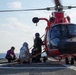 A red helicopter, consistent with a U.S. Coast Guard MH-65 Dolphin, is landed on the flight deck of a naval vessel at sea. Several personnel in specialized gear are working around the aircraft, with one individual appearing to be on a stretcher or litter, suggesting a medical evacuation or rescue operation. The rotor blades are spinning, indicating the helicopter is active.