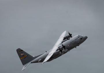 A U.S. Air Force C-130 Hercules turboprop military transport aircraft is captured in flight against an overcast gray sky. The aircraft is in a climbing or banking attitude, displaying standard gray military livery with visible U.S. Air Force markings and an American flag on the tail. The image appears to have been taken during an airshow or training flight.