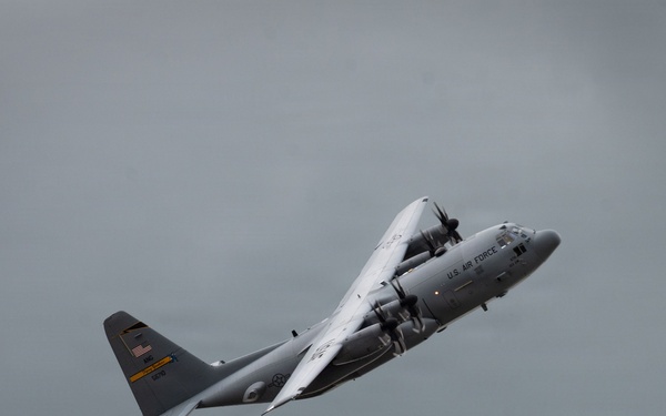 A U.S. Air Force C-130 Hercules turboprop military transport aircraft is captured in flight against an overcast gray sky. The aircraft is in a climbing or banking attitude, displaying standard gray military livery with visible U.S. Air Force markings and an American flag on the tail. The image appears to have been taken during an airshow or training flight.
