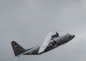A U.S. Air Force C-130 Hercules military transport aircraft is captured in flight against an overcast, gray sky. The aircraft is in a banking maneuver, showing its underside and port side. The plane bears standard U.S. Air Force gray livery with visible markings including an American flag on the tail.