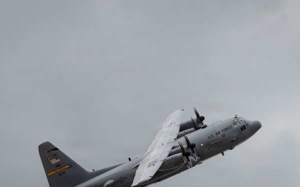 A U.S. Air Force C-130 Hercules military transport aircraft is captured in flight against an overcast, gray sky. The aircraft is in a banking maneuver, showing its underside and port side. The plane bears standard U.S. Air Force gray livery with visible markings including an American flag on the tail.