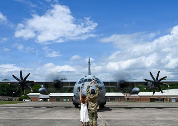 A military service member in camouflage uniform and a civilian in a white dress stand together on an airfield tarmac with their arms raised, facing a large military turboprop aircraft head-on. The aircraft, identifiable as a C-130 Hercules, has its four propellers spinning. The scene appears to capture a departure or arrival ceremony at a military air base.
