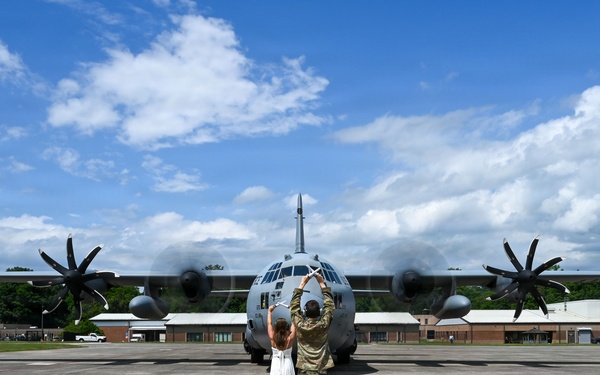 A military service member in camouflage uniform and a civilian in a white dress stand together on an airfield tarmac with their arms raised, facing a large military turboprop aircraft head-on. The aircraft, identifiable as a C-130 Hercules, has its four propellers spinning. The scene appears to capture a departure or arrival ceremony at a military air base.