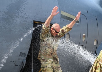 A military service member in OCP (Operational Camouflage Pattern) uniform is being doused with water near the door of a large military aircraft, likely a C-130 or similar transport. This appears to be a traditional military 'fini flight' water dousing ceremony, marking a final flight milestone. A second uniformed service member observes from the right side of the frame.