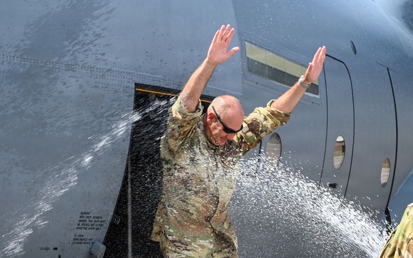 A military service member in OCP (Operational Camouflage Pattern) uniform is being doused with water near the door of a large military aircraft, likely a C-130 or similar transport. This appears to be a traditional military 'fini flight' water dousing ceremony, marking a final flight milestone. A second uniformed service member observes from the right side of the frame.
