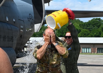A military celebration scene at a Connecticut Air National Guard base, where a service member in camouflage uniform is being doused with water from a yellow Igloo cooler by a smiling colleague in a flight suit. A third person in the foreground appears to be spraying something from a green bottle. This type of water dousing is a traditional military 'fini flight' or retirement/milestone celebration ritual.