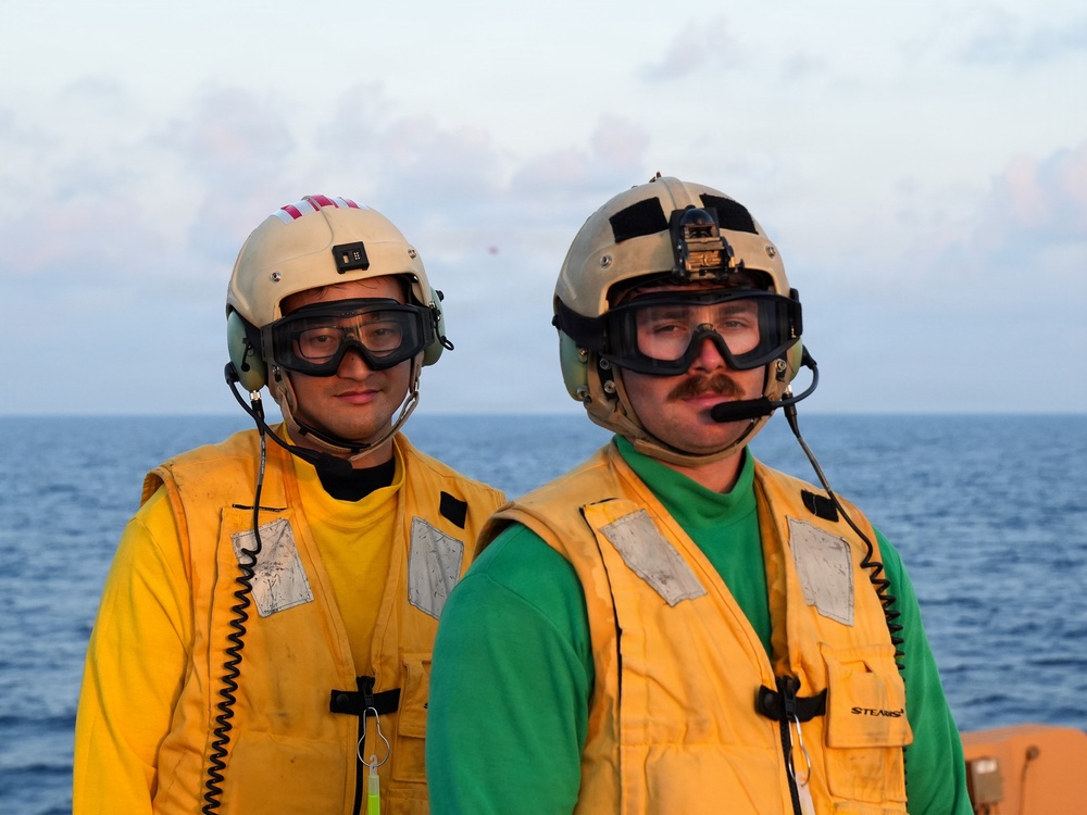 Two military personnel are standing on what appears to be a naval vessel deck with open ocean visible in the background. Both individuals are wearing yellow safety vests, protective goggles, helmets with communication headsets, and microphones, indicating they are engaged in flight deck or maritime operations. The image was likely taken during dawn or dusk based on the soft lighting conditions.